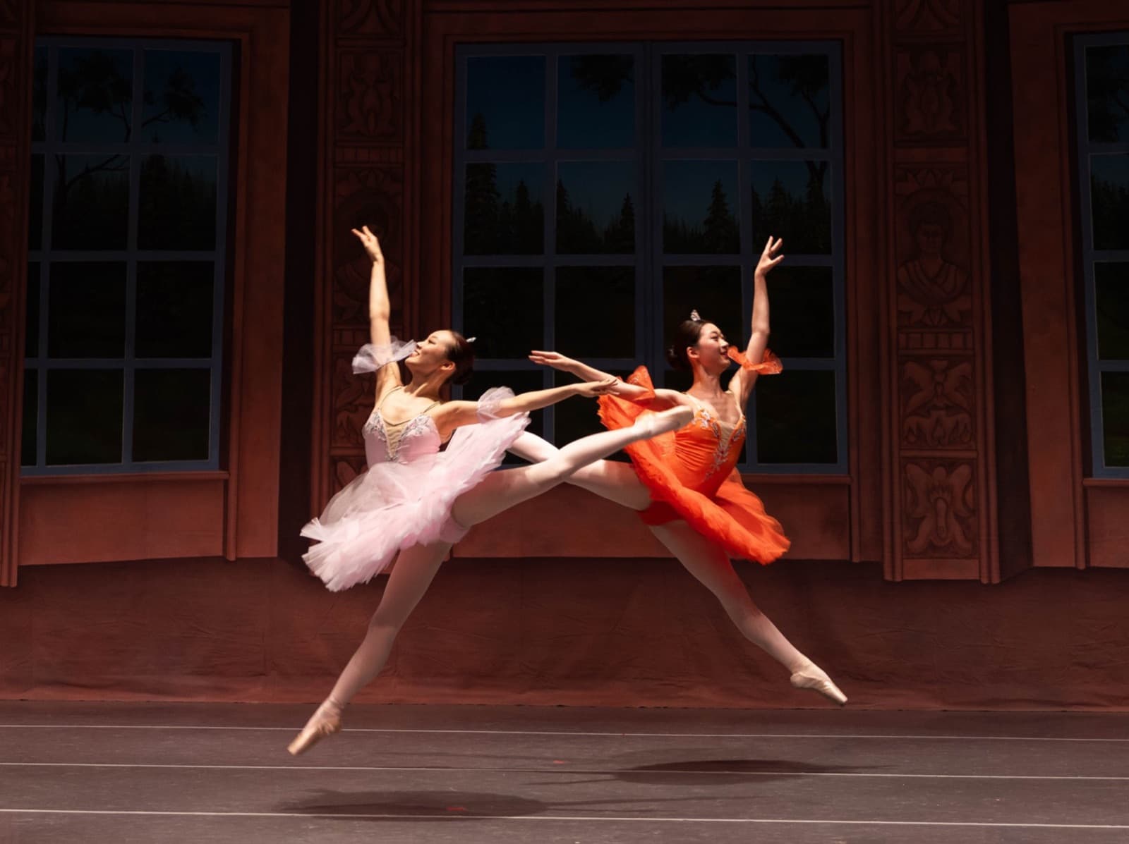 Two ballerinas, one in a pink tutu and one in an orange tutu, leaping side by side in a grand jeté before a painted window backdrop.